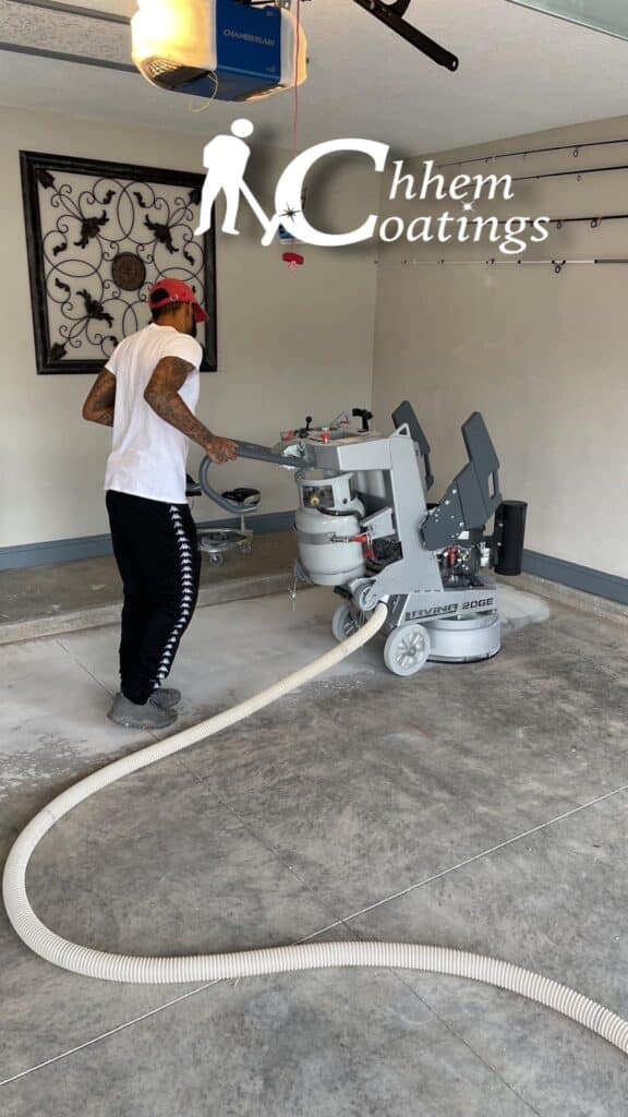 A person operates a floor grinding machine in a garage with light-colored walls, equipped with a decorated iron wall piece. The atmosphere appears industrious.